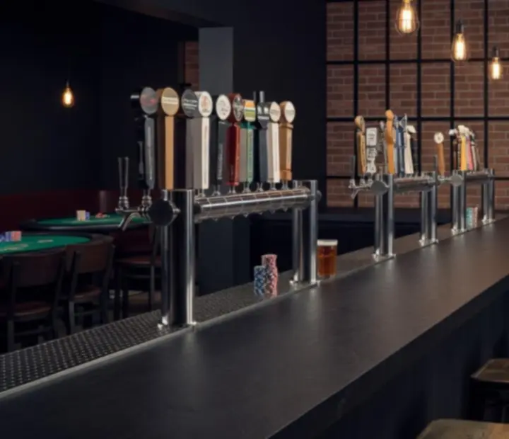 Industrial taproom bar with exposed steel, taps, and a glimpse of the card lounge.